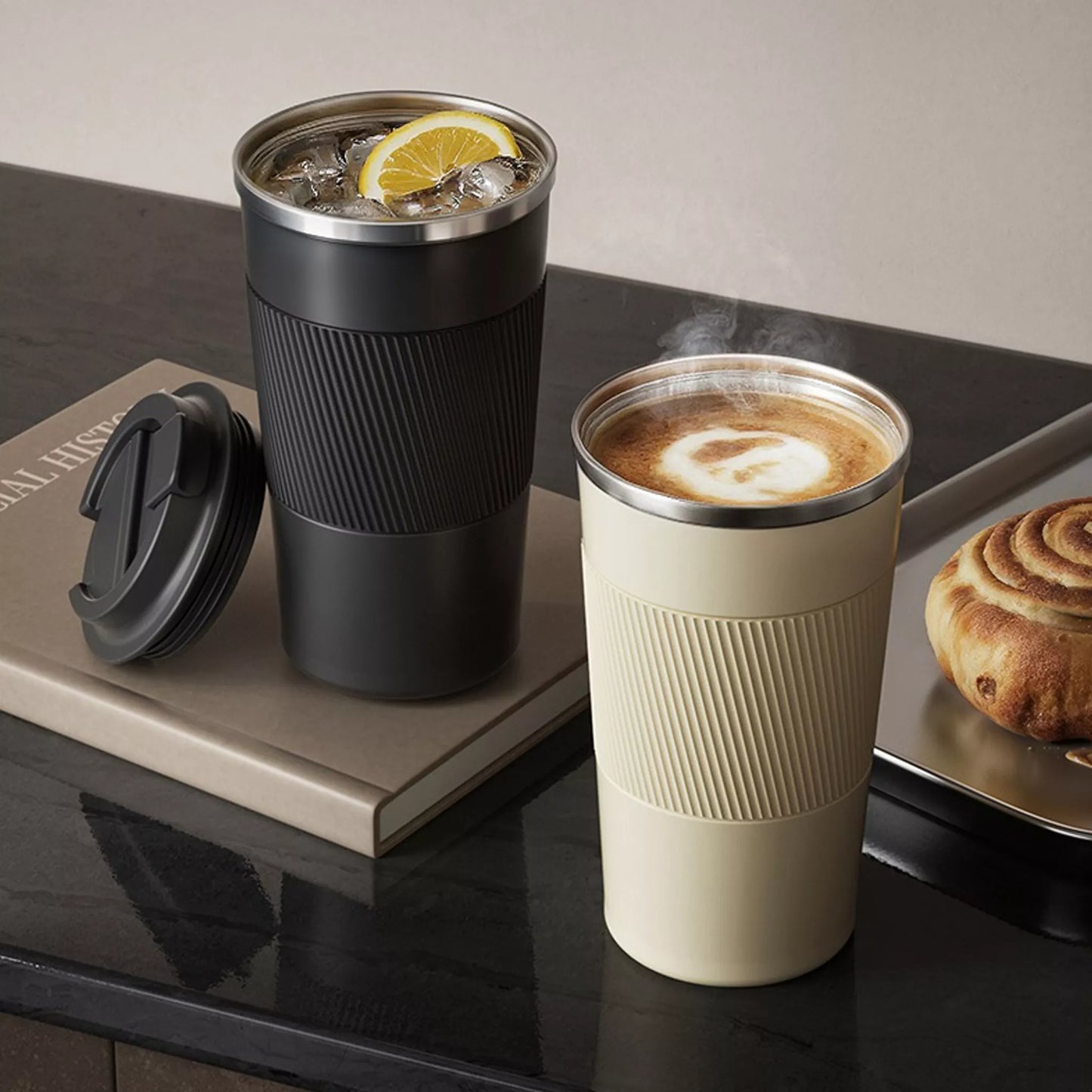 Two travel mugs, one black and one beige, on a reflective surface with a book and pastry in the background.