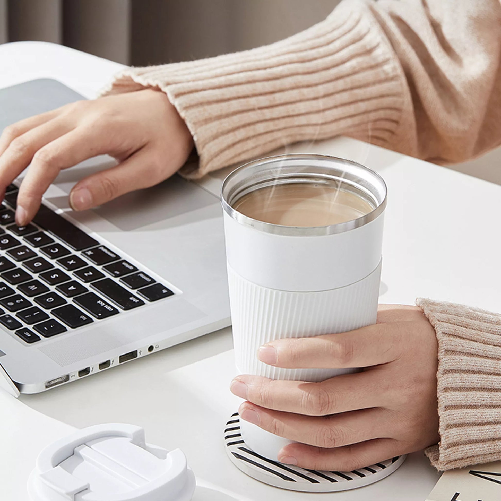 Person holding a steaming cup of coffee next to a laptop on a desk.
